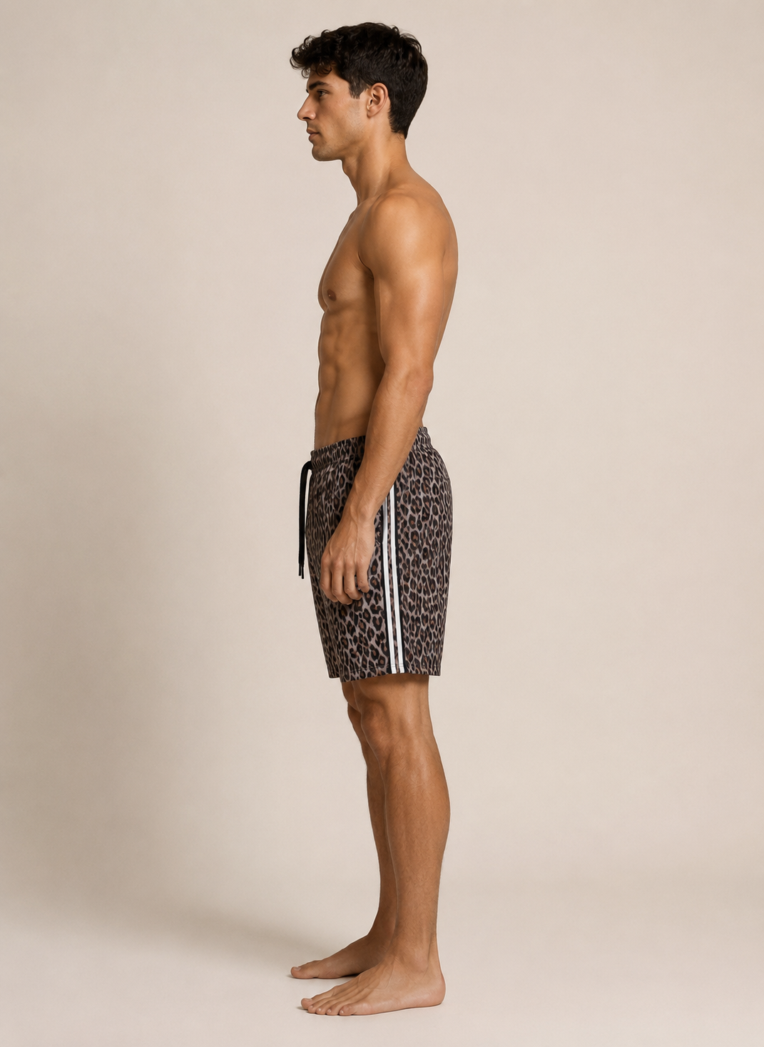 A man shown in profile wearing patterned swim trunks with white side stripes, standing barefoot against a plain beige studio background.