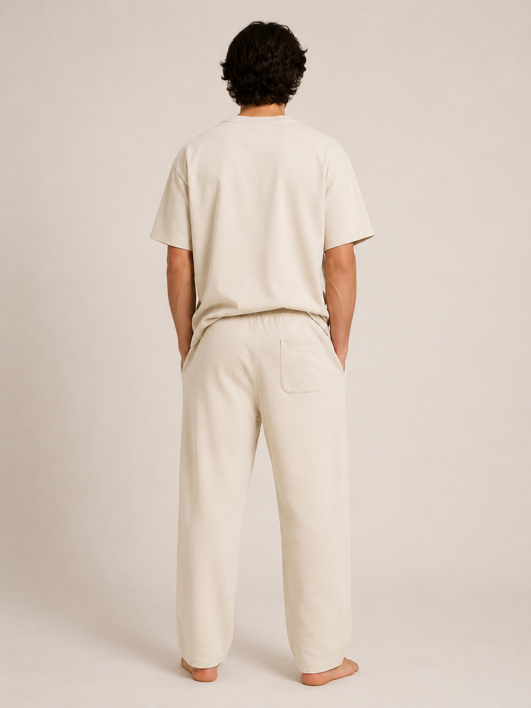 A man viewed from behind wearing a light cream-colored short-sleeve t-shirt and matching wide-leg lounge pants, standing barefoot against a plain beige studio background.