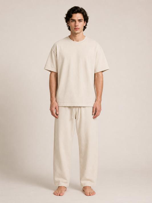 A man stands barefoot against a plain warm beige studio background, wearing a loose short-sleeve cream T-shirt and matching cream drawstring pants.