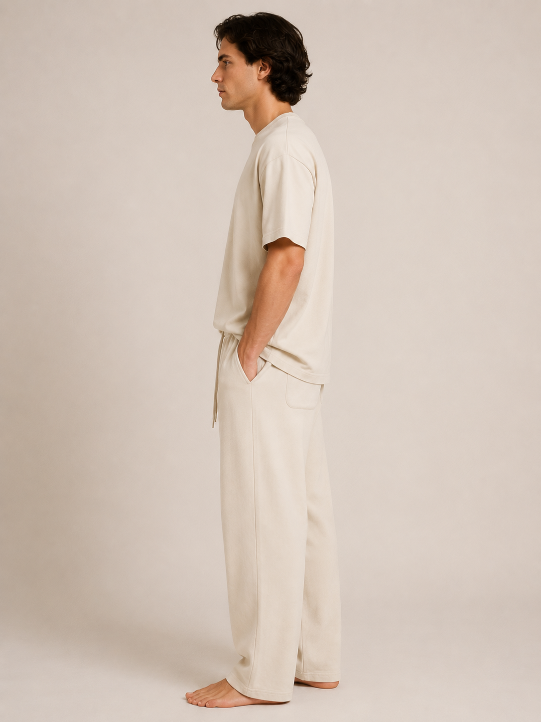 A man in a side profile wearing a matching cream-colored short-sleeve t-shirt and wide-leg trousers, standing barefoot against a solid light beige studio background.