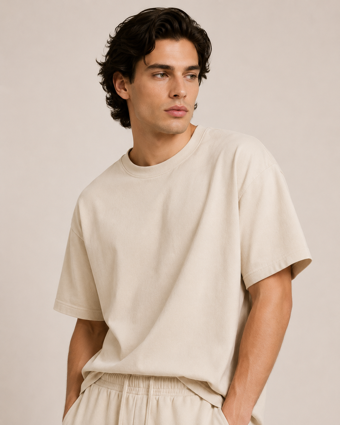 A man wearing a relaxed-fit, short-sleeve crewneck t-shirt in a light beige or cream color, paired with matching light-colored drawstring pants, set against a solid light beige studio background.