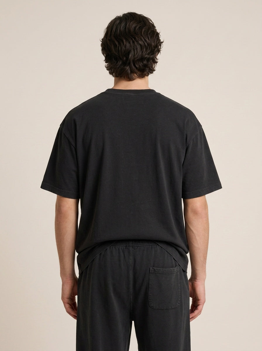 A man viewed from the back wearing a black short-sleeve t-shirt and black pants against a plain beige studio background.