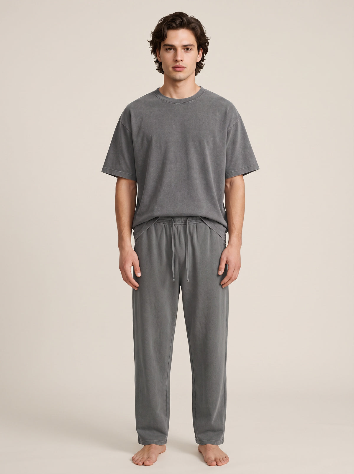 A man stands barefoot against a plain warm beige studio background, wearing a loose gray short-sleeve T-shirt and matching gray drawstring pants.