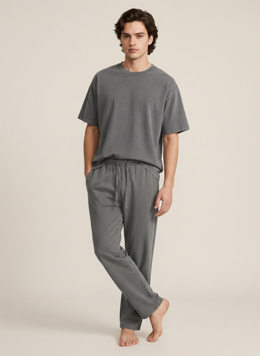 A man modeling a matching gray lounge set consisting of a short-sleeve crewneck t-shirt and drawstring sweatpants, photographed against a solid light beige studio background.