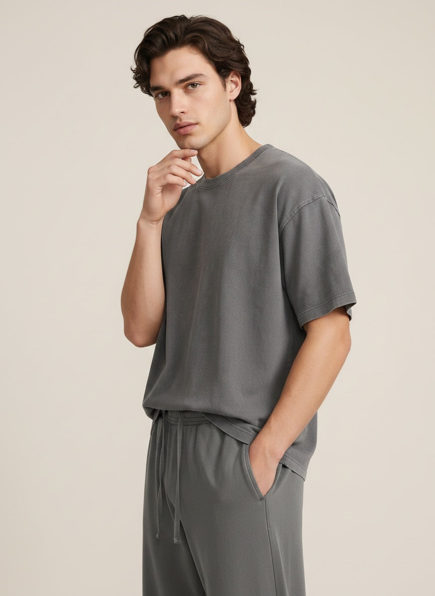 A man wearing a loose-fit gray short-sleeve t-shirt and matching gray drawstring pants, standing against a plain beige studio background.