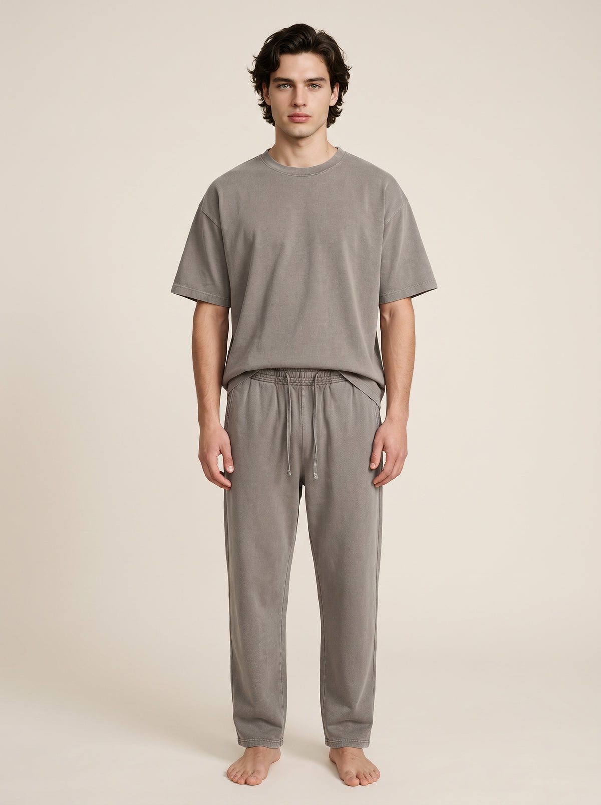 A man stands barefoot against a plain warm beige studio background, wearing a matching gray lounge set consisting of a short-sleeve crew-neck t-shirt and drawstring sweatpants.