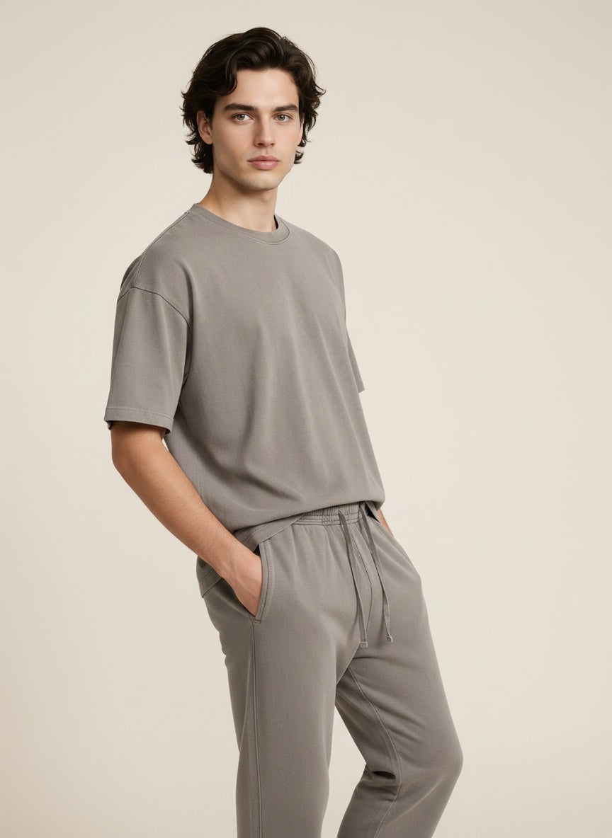 A man wearing a light gray short-sleeve crewneck t-shirt and matching gray drawstring sweatpants, standing against a solid light beige background.
