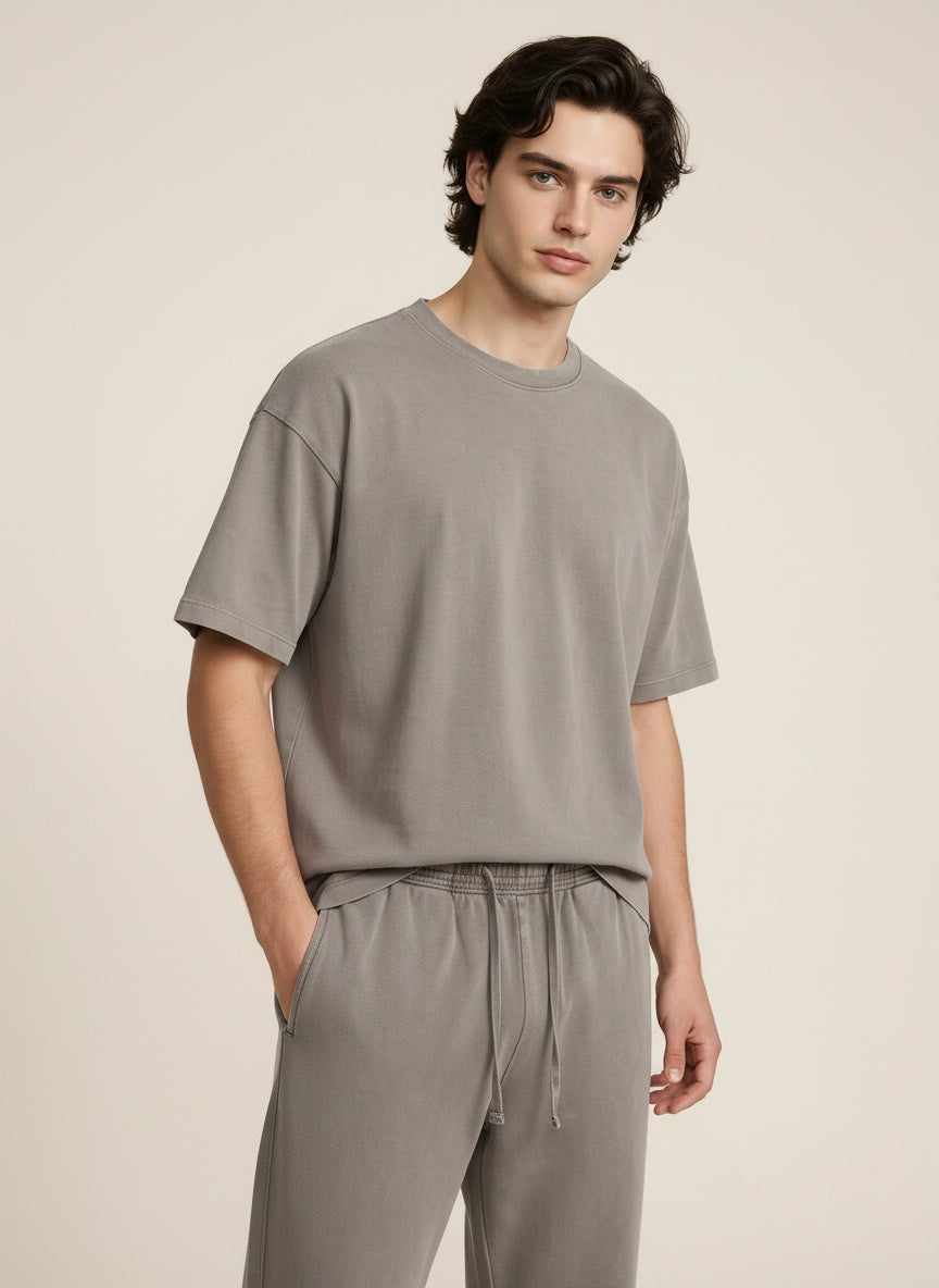 A man wearing a light gray short-sleeve crewneck t-shirt and matching gray drawstring sweatpants, standing against a solid beige studio background.