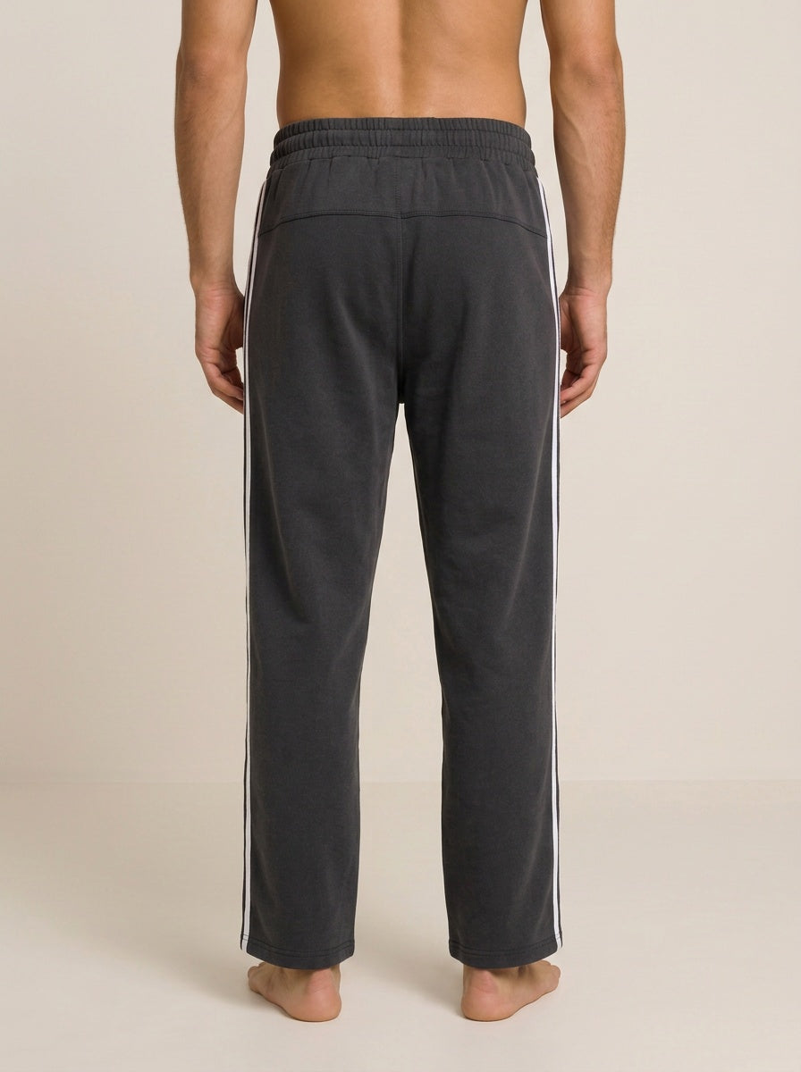 A man viewed from the back wearing dark charcoal gray track pants with white side stripes, standing barefoot against a plain beige studio background.