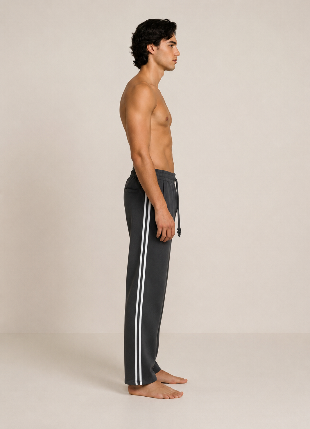 A man stands in profile wearing dark charcoal gray track pants with two white side stripes and a drawstring waist, photographed against a plain warm off-white studio background.