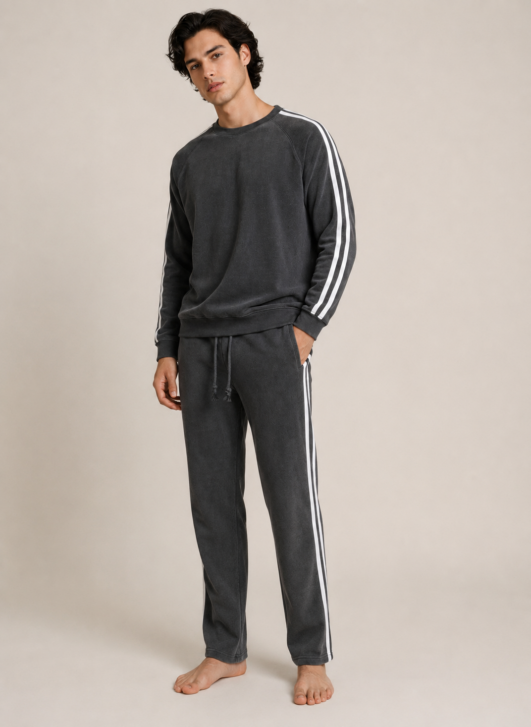 A man modeling a dark gray two-piece loungewear set with white three-stripe detailing on the sleeves and pants, standing against a plain beige studio background.