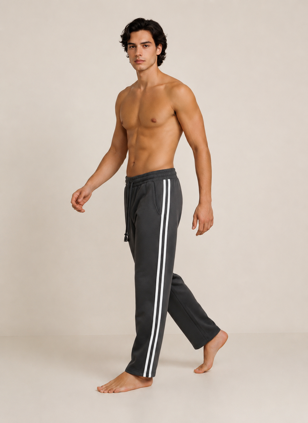 A shirtless male model walking in profile wearing dark charcoal gray track pants with white side stripes, against a plain beige studio background.