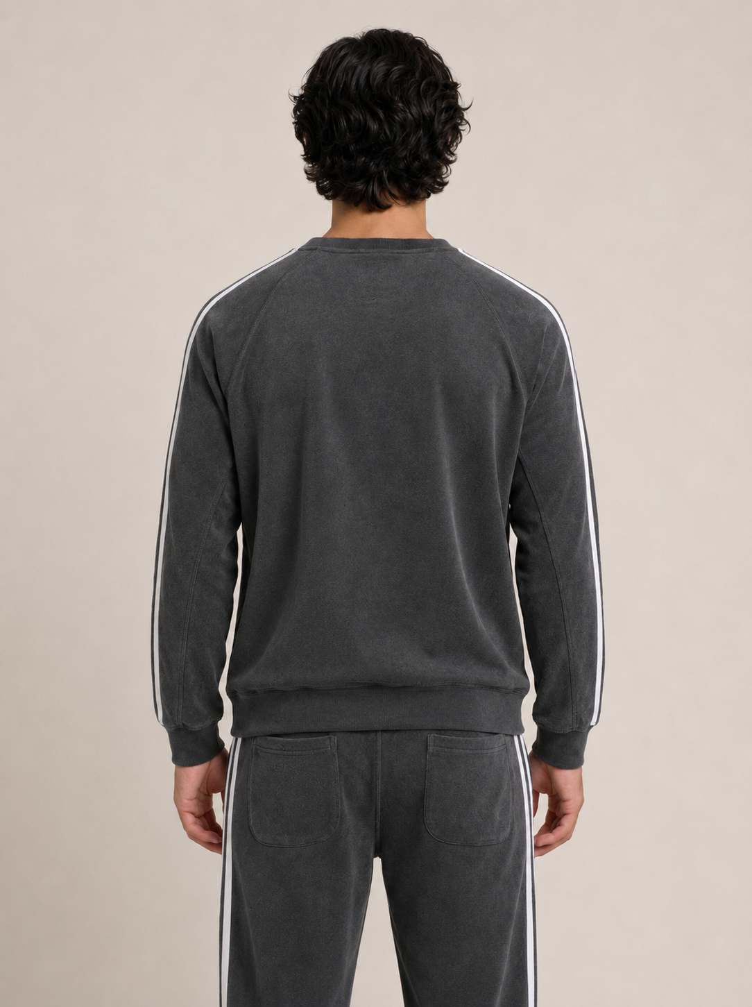 A man viewed from the back wearing a dark gray crewneck sweatshirt and matching sweatpants, both featuring white stripes down the sleeves and side seams, against a solid beige background.