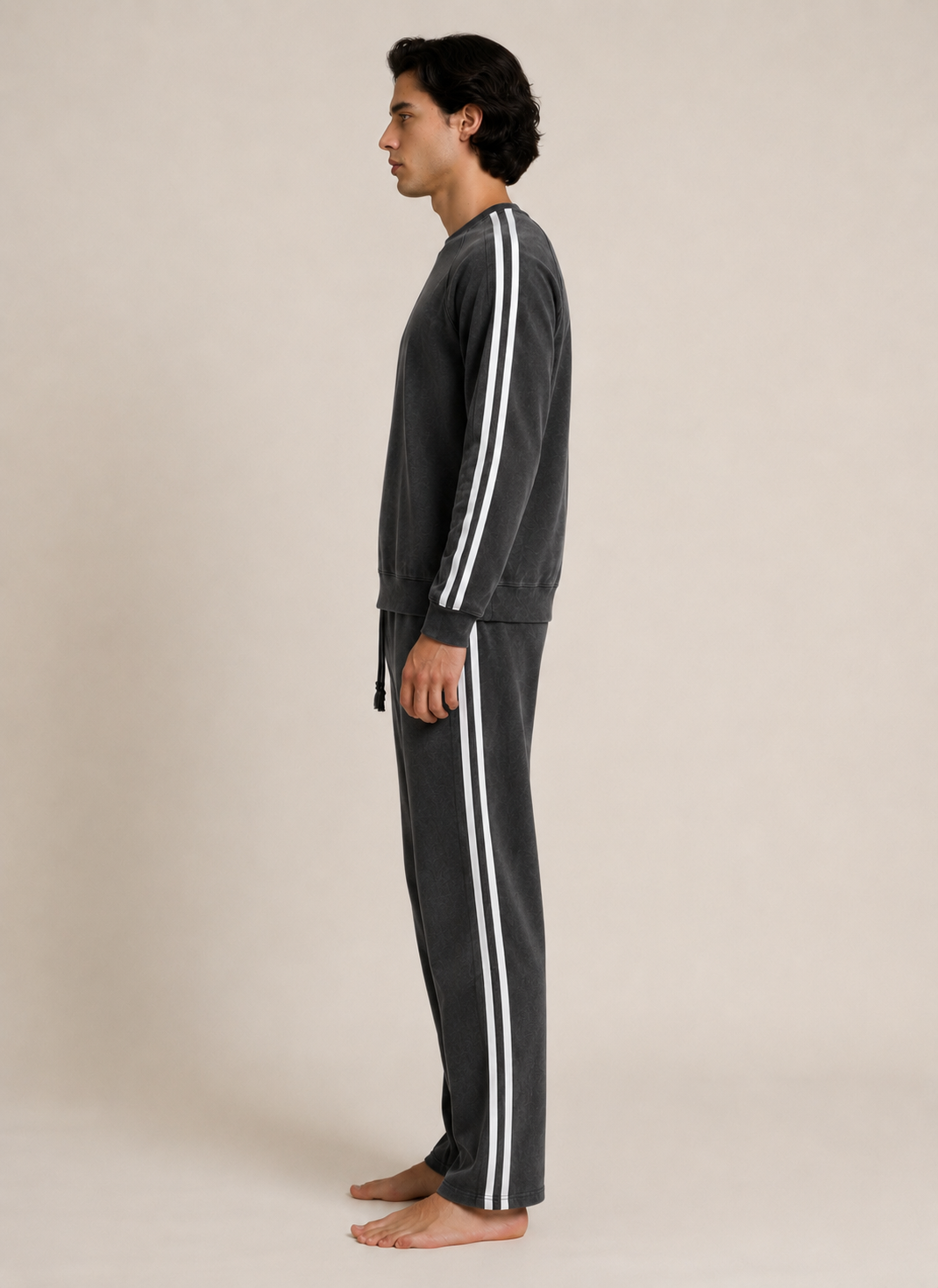 A man in profile wearing a dark gray two-piece tracksuit with white double stripes down the sleeves and legs, standing barefoot against a plain beige studio background.