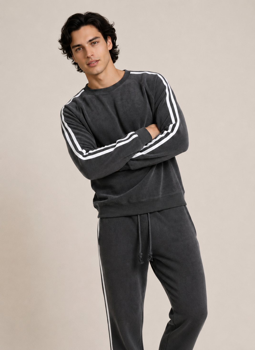 A man modeling a dark gray sweatshirt and matching sweatpants set. The sweatshirt features white stripes down the sleeves, and the sweatpants have white side stripes, set against a plain beige studio background.