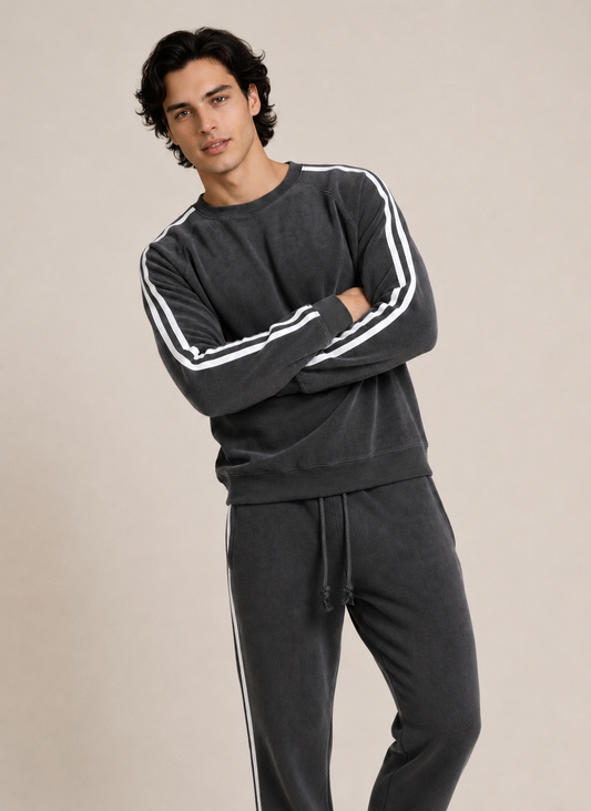 A man modeling a dark gray sweatshirt and matching sweatpants set. The sweatshirt features white stripes down the sleeves, and the sweatpants have white side stripes, set against a plain beige studio background.