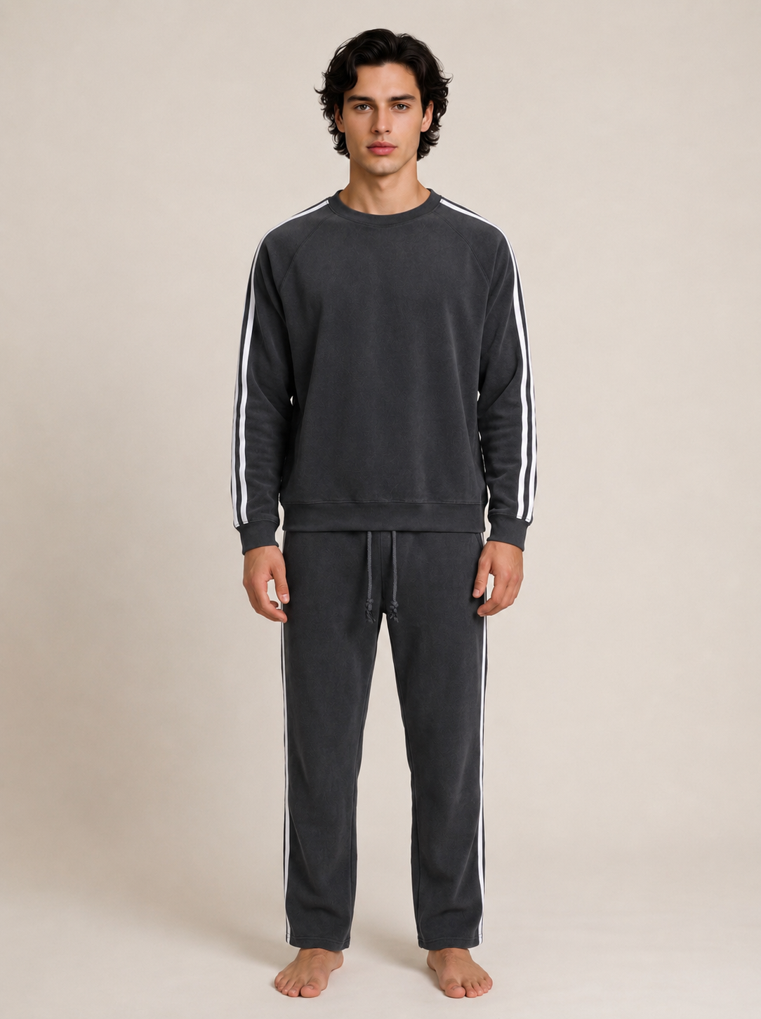 A man modeling a dark gray sweatshirt and matching sweatpants set with white stripes down the sleeves and sides, standing against a plain beige studio background.