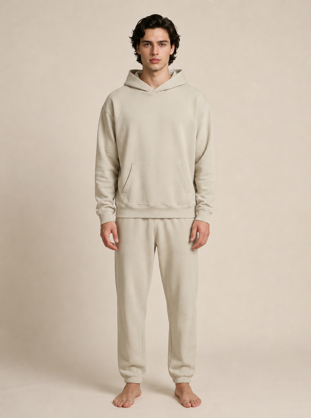 A man stands barefoot wearing a matching light beige hooded sweatshirt and jogger sweatpants set against a plain beige studio background.