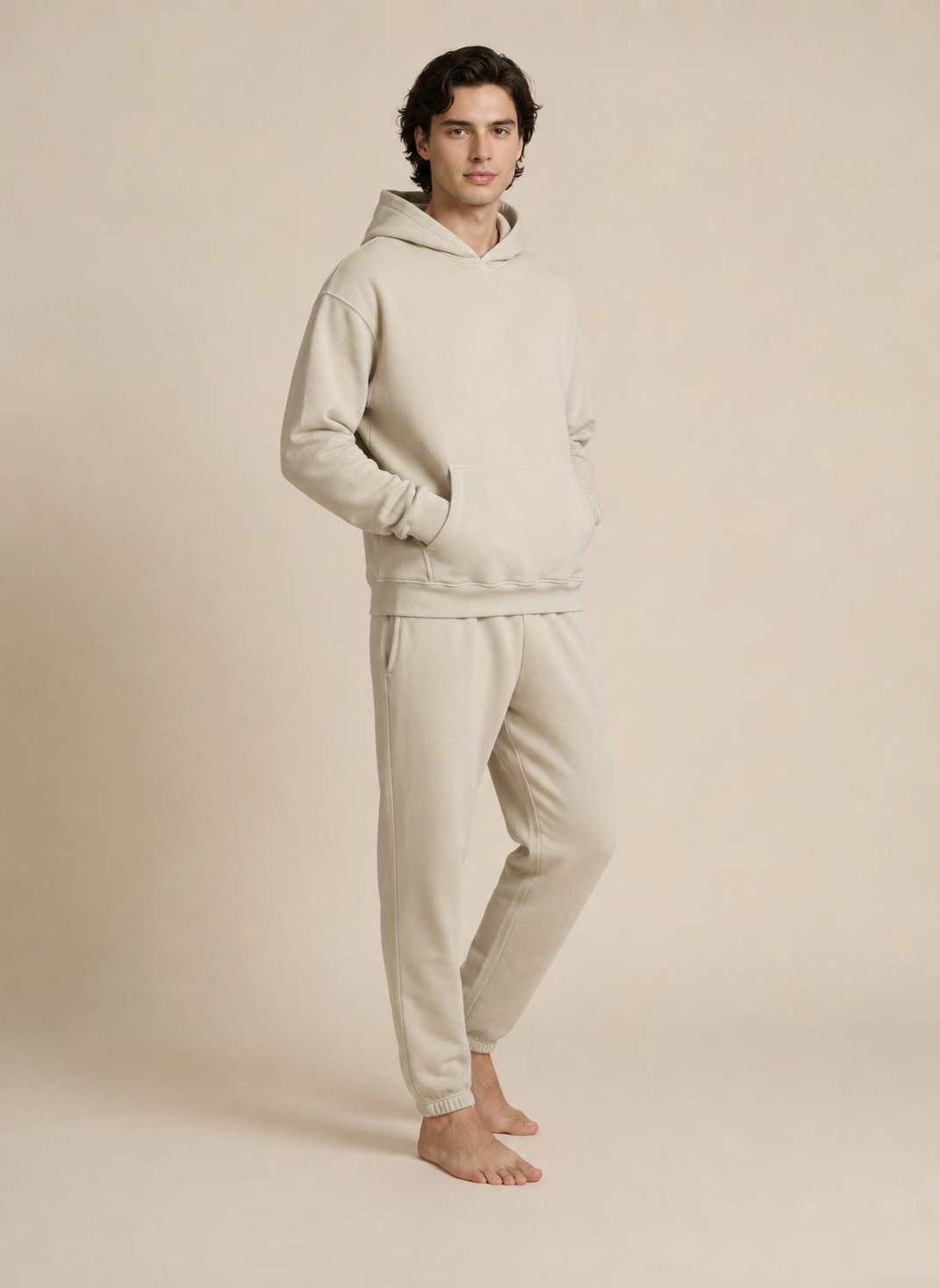 A man modeling a matching light beige hoodie and sweatpants lounge set against a solid beige studio background.