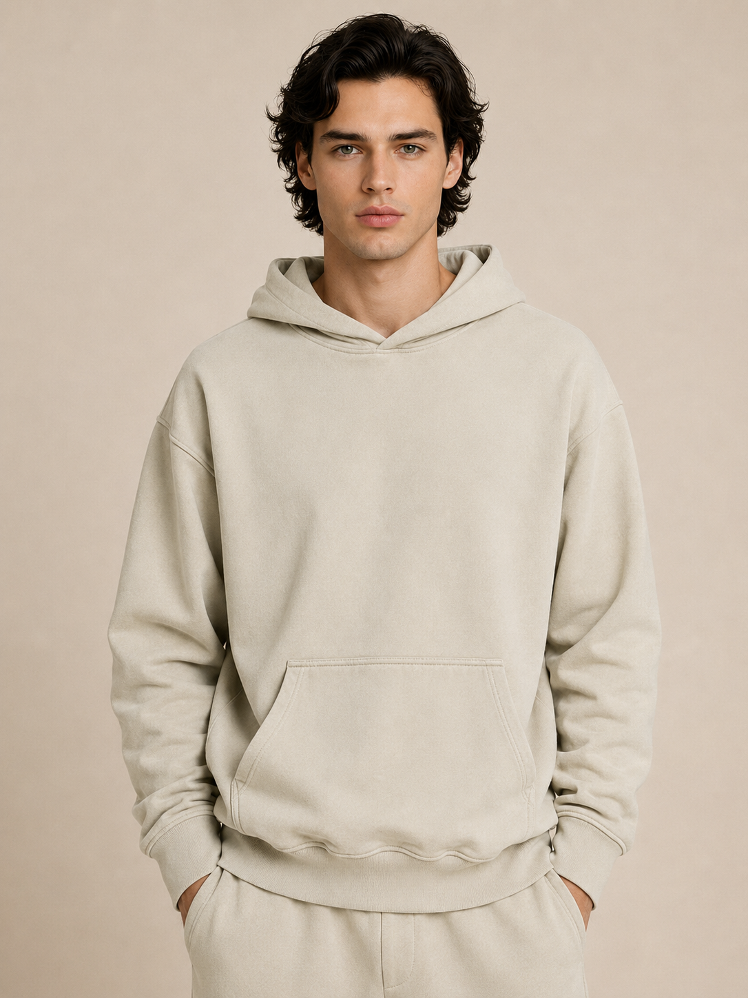 A man wearing a light beige pullover hoodie and matching sweatpants, standing against a solid beige studio background.