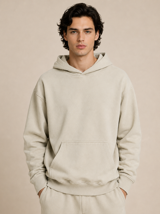 A man wearing a light beige pullover hoodie and matching sweatpants, standing against a solid beige studio background.
