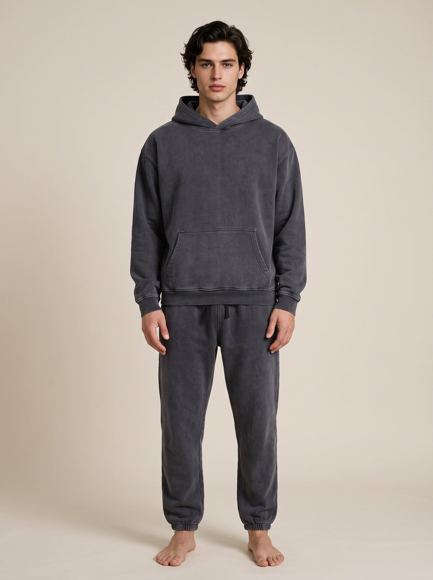 A man wearing a dark gray two-piece loungewear set consisting of a hooded sweatshirt and matching sweatpants, standing against a solid beige background.