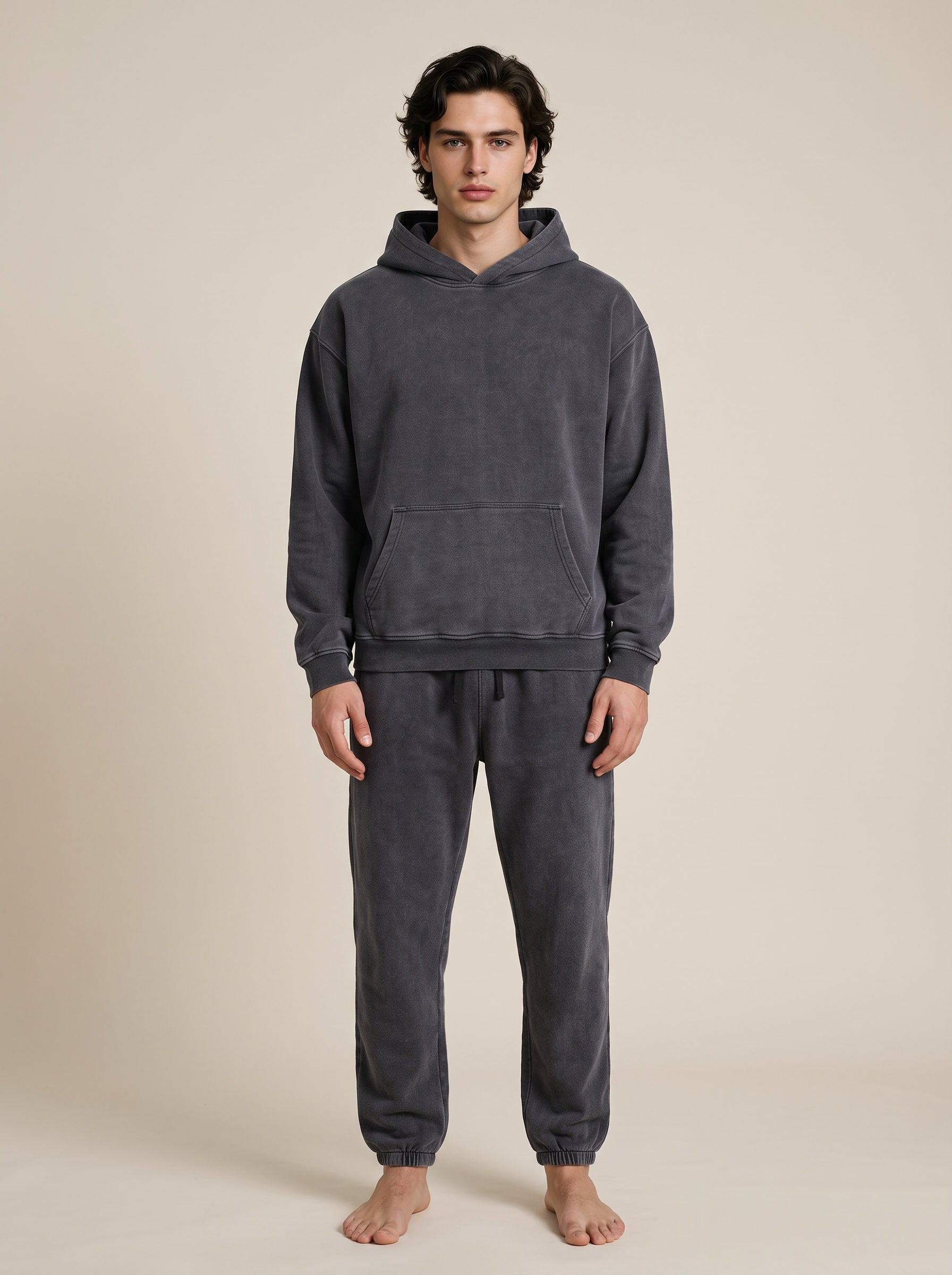 A man wearing a dark gray two-piece loungewear set consisting of a hooded sweatshirt and matching sweatpants, standing against a solid beige background.