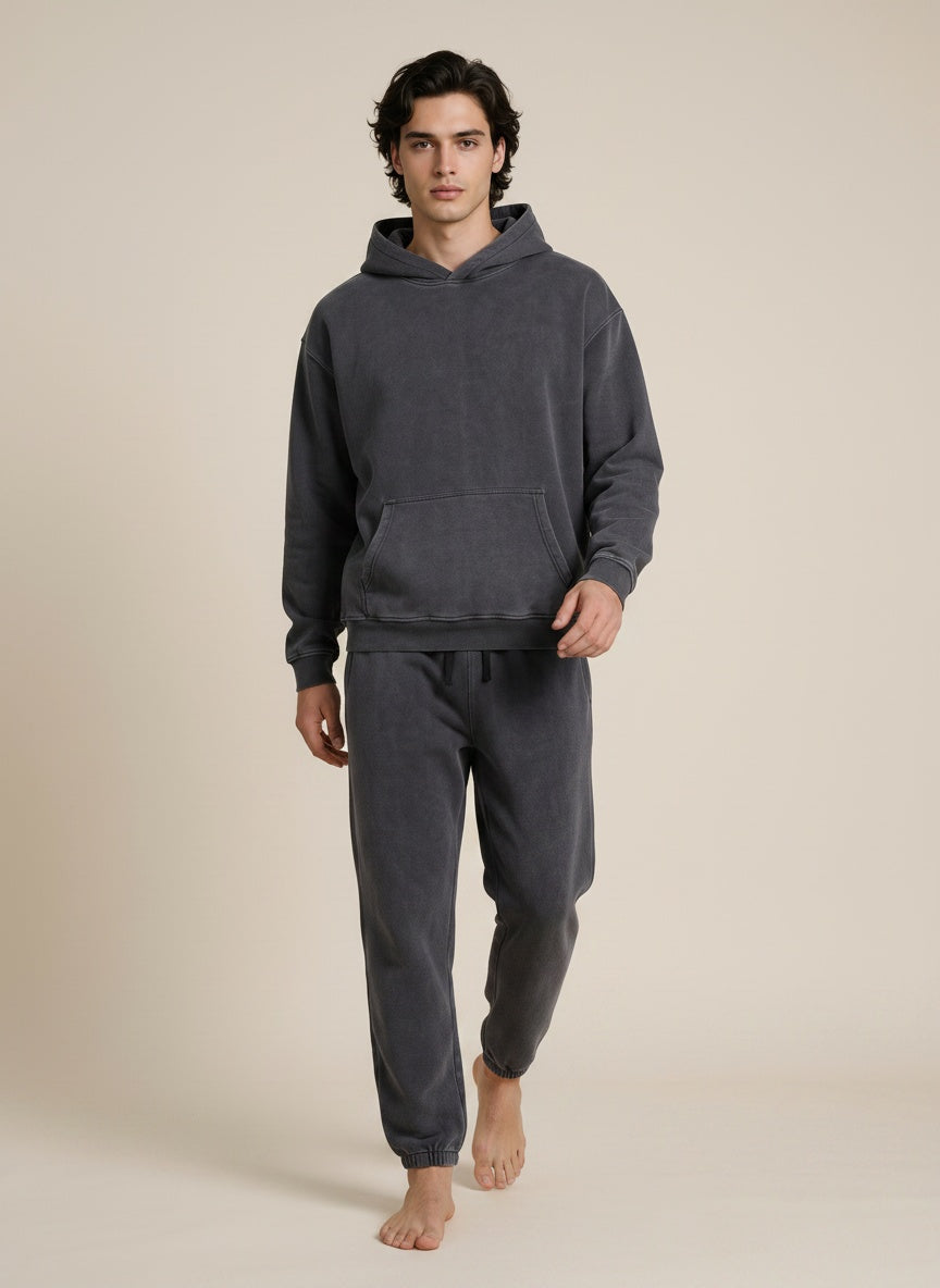 A man modeling a dark gray two-piece loungewear set consisting of a pullover hoodie and matching jogger pants, standing against a solid beige studio background.