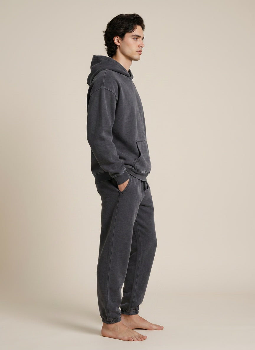 A man wearing a dark gray matching lounge set consisting of a hooded sweatshirt and jogger pants, standing in profile against a solid light beige background.