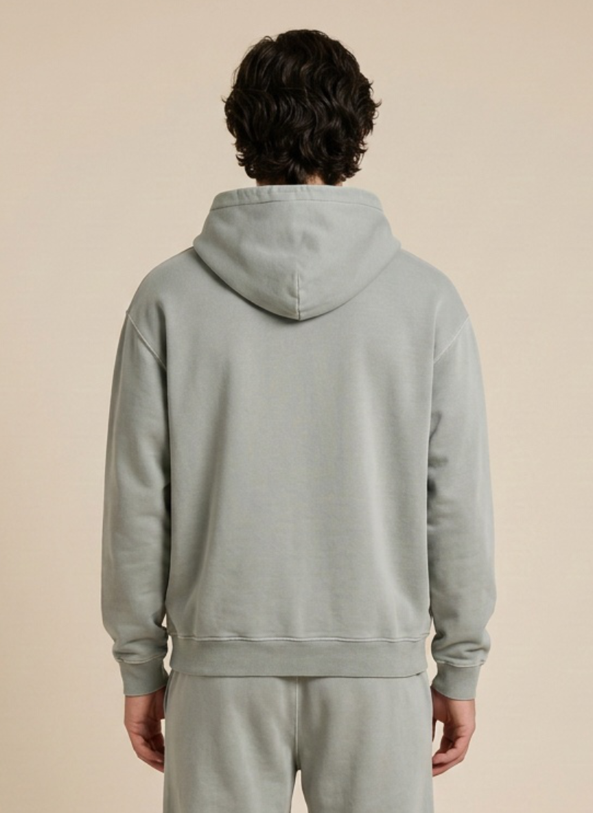 A person viewed from the back wearing a light sage green hooded sweatshirt and matching sweatpants against a plain beige studio background.