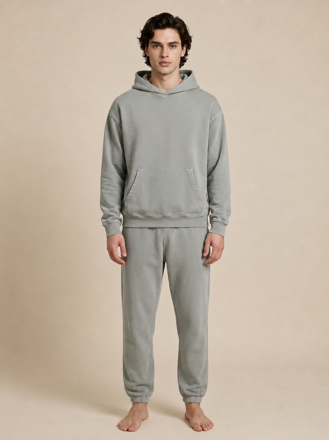 A man wearing a matching light gray fleece hoodie and jogger sweatpants set, standing barefoot against a solid beige studio background.