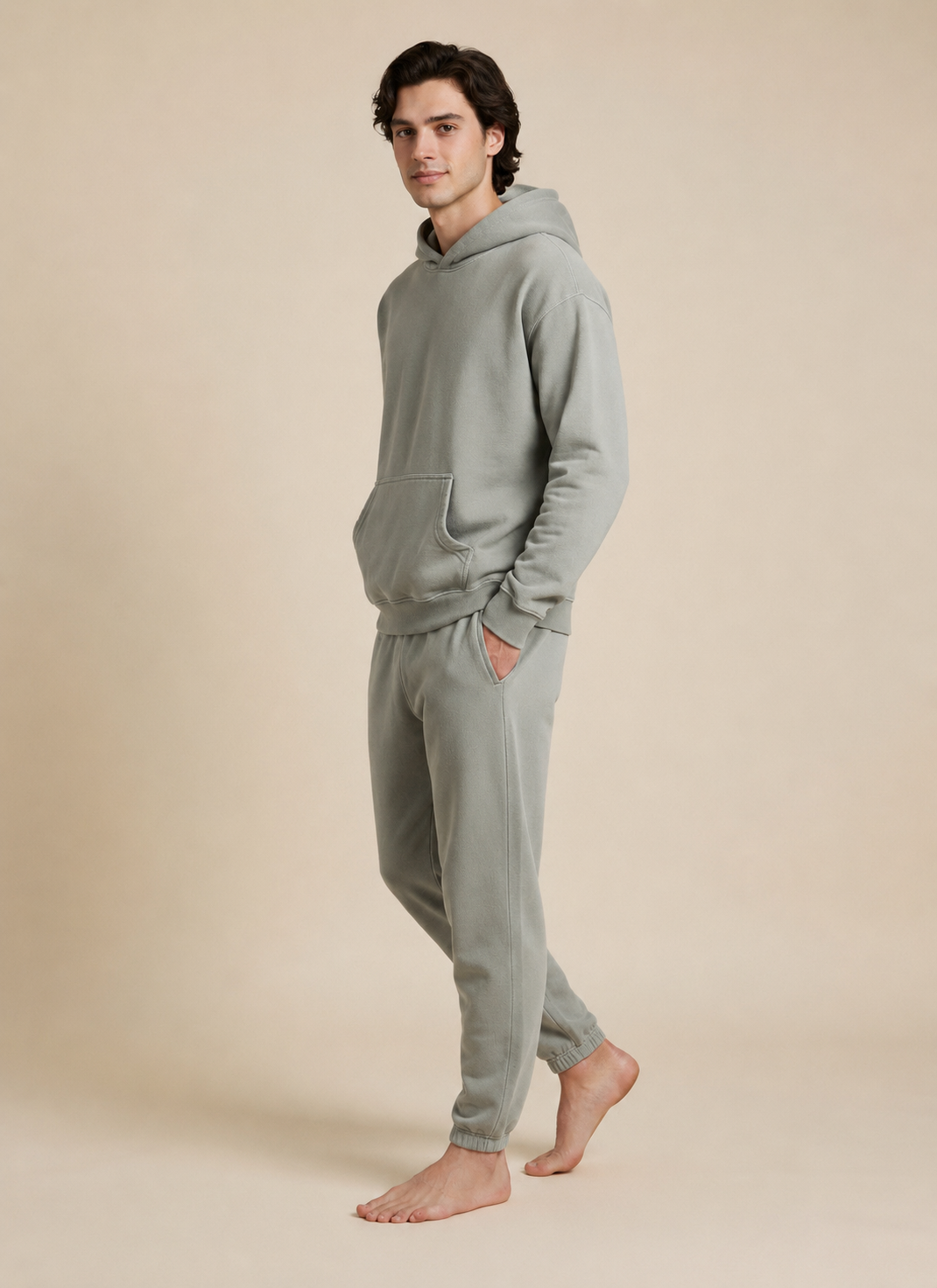 A man wearing a matching sage green fleece hoodie and jogger sweatpants set, standing barefoot against a solid beige studio background.