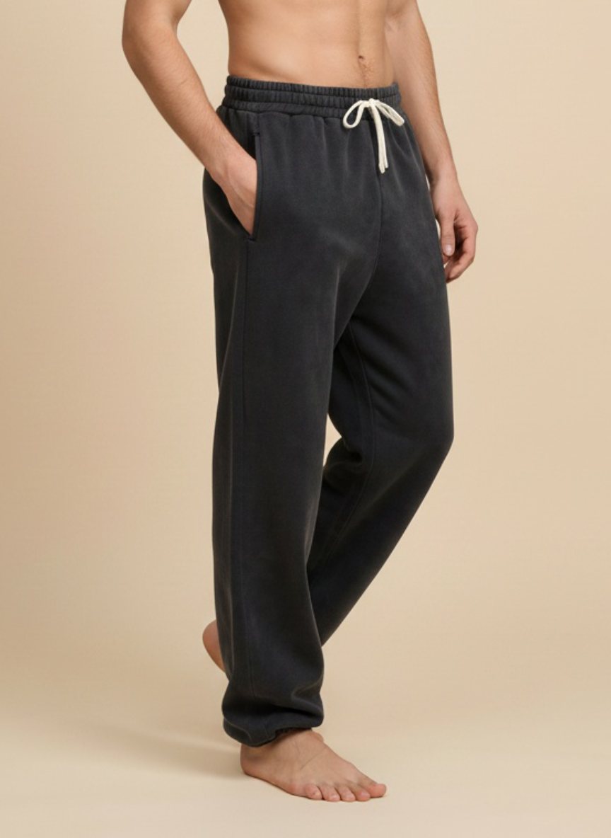 A man wearing dark charcoal gray lounge sweatpants with a white drawstring, standing barefoot against a solid beige background.