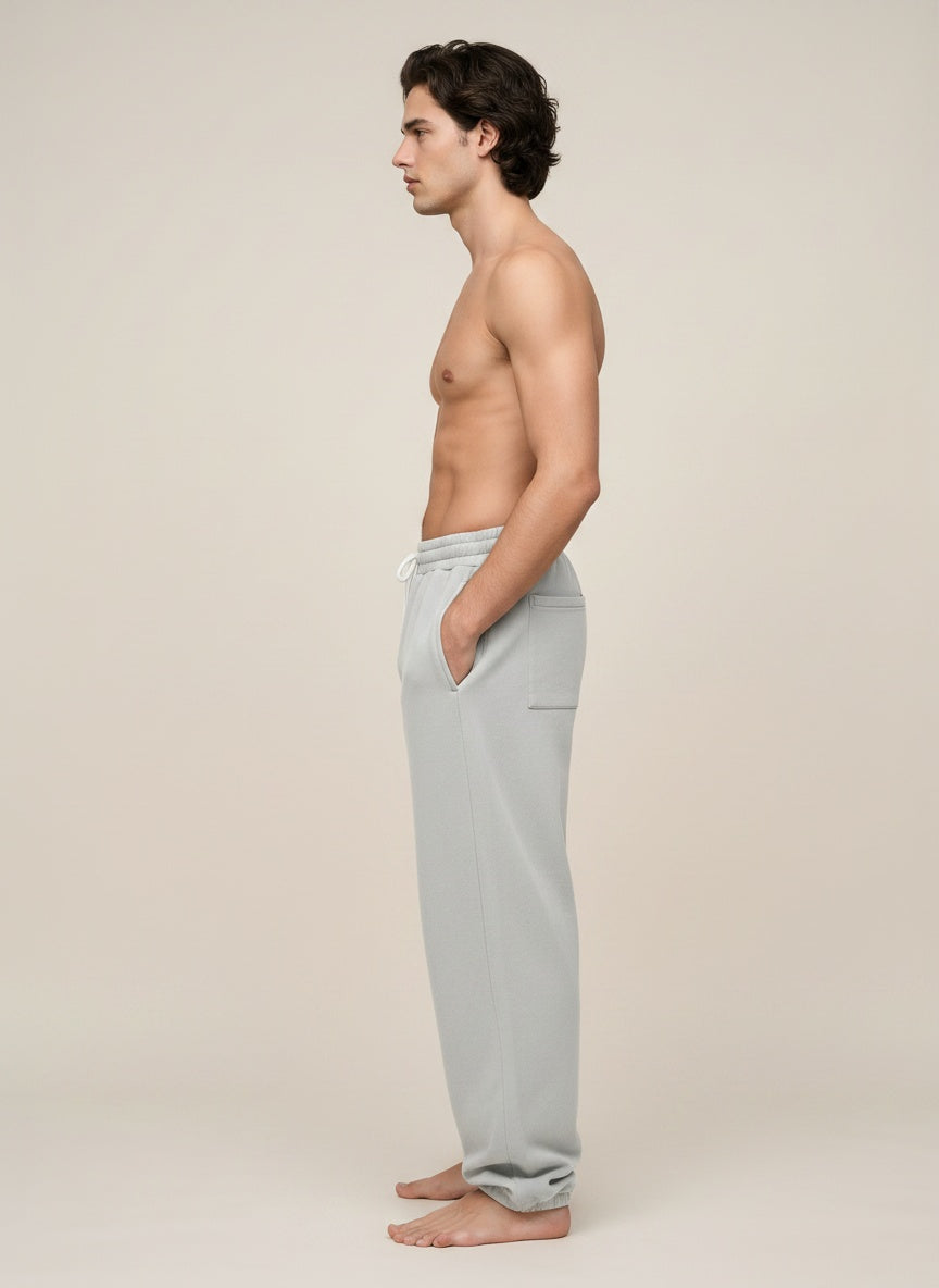 A man in side profile wearing light gray lounge pants, standing barefoot against a plain beige studio background.