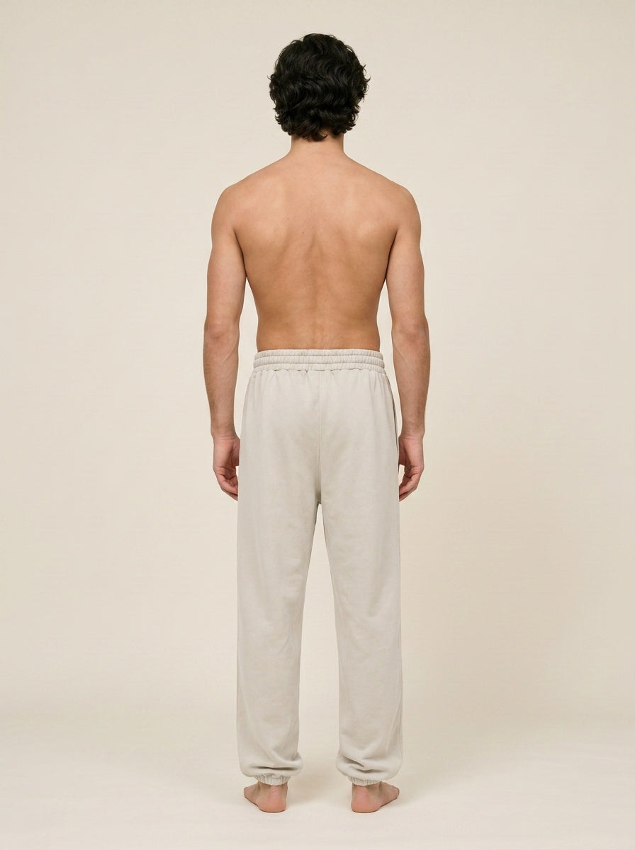 A man viewed from behind wearing light beige lounge pants, standing barefoot against a plain beige studio background.