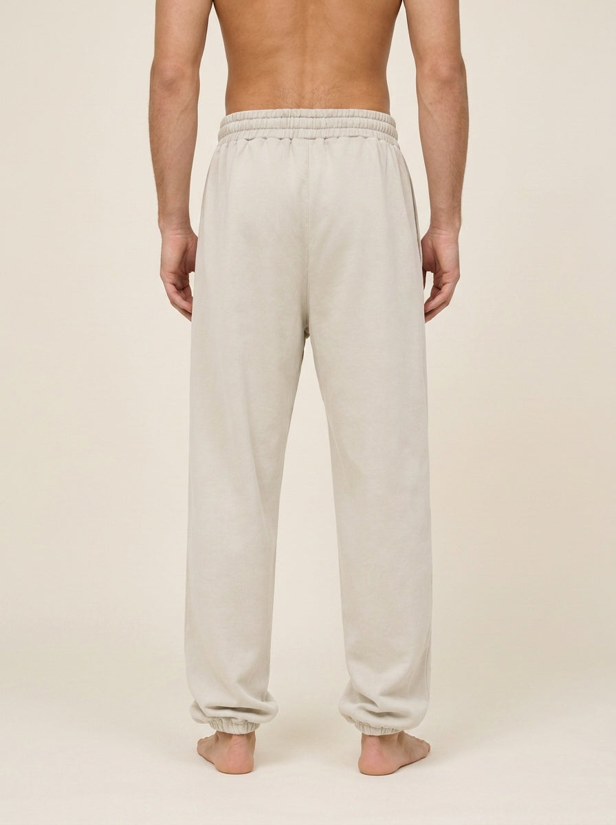 A man viewed from the back wearing light beige sweatpants with an elastic waistband and cuffs, standing barefoot against a plain cream studio background.