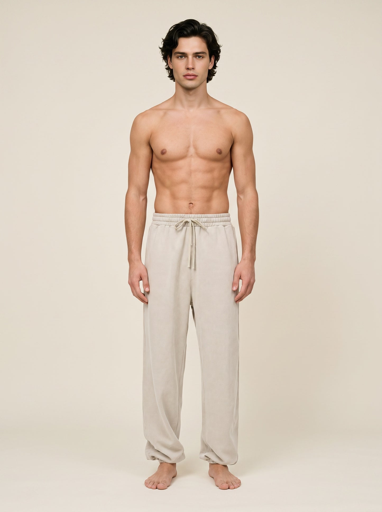 A shirtless male model stands barefoot against a plain warm beige studio background, wearing loose light beige drawstring lounge pants.