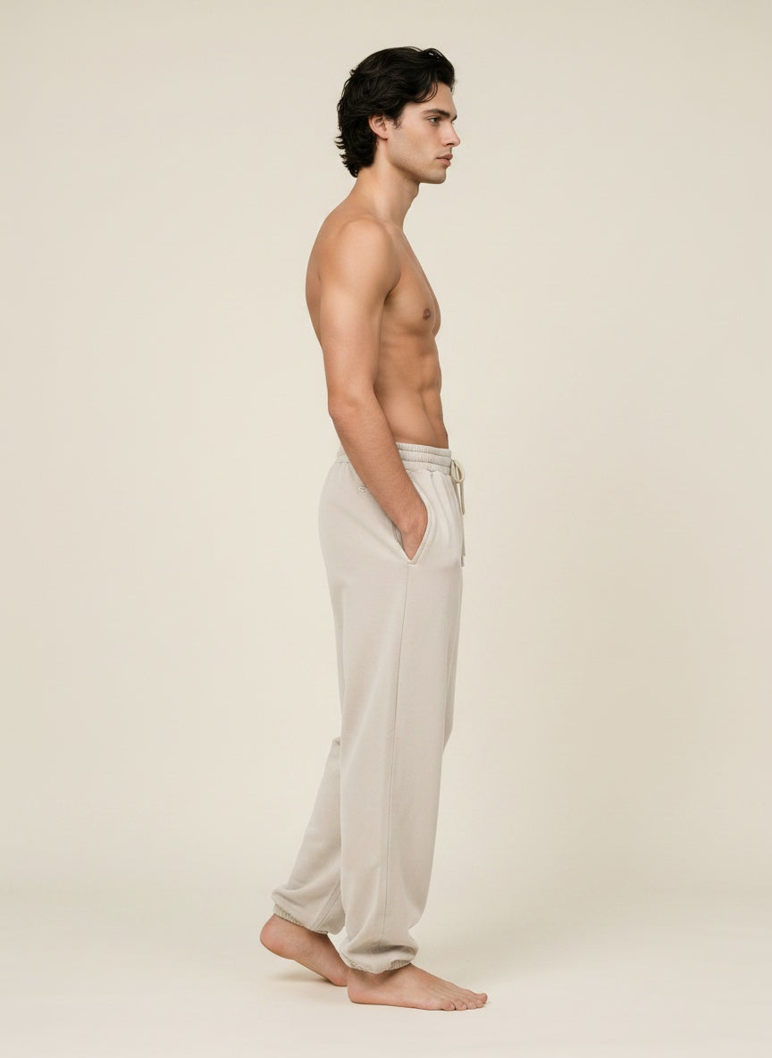 A man in side profile wearing light beige drawstring sweatpants, standing barefoot against a plain beige studio background.