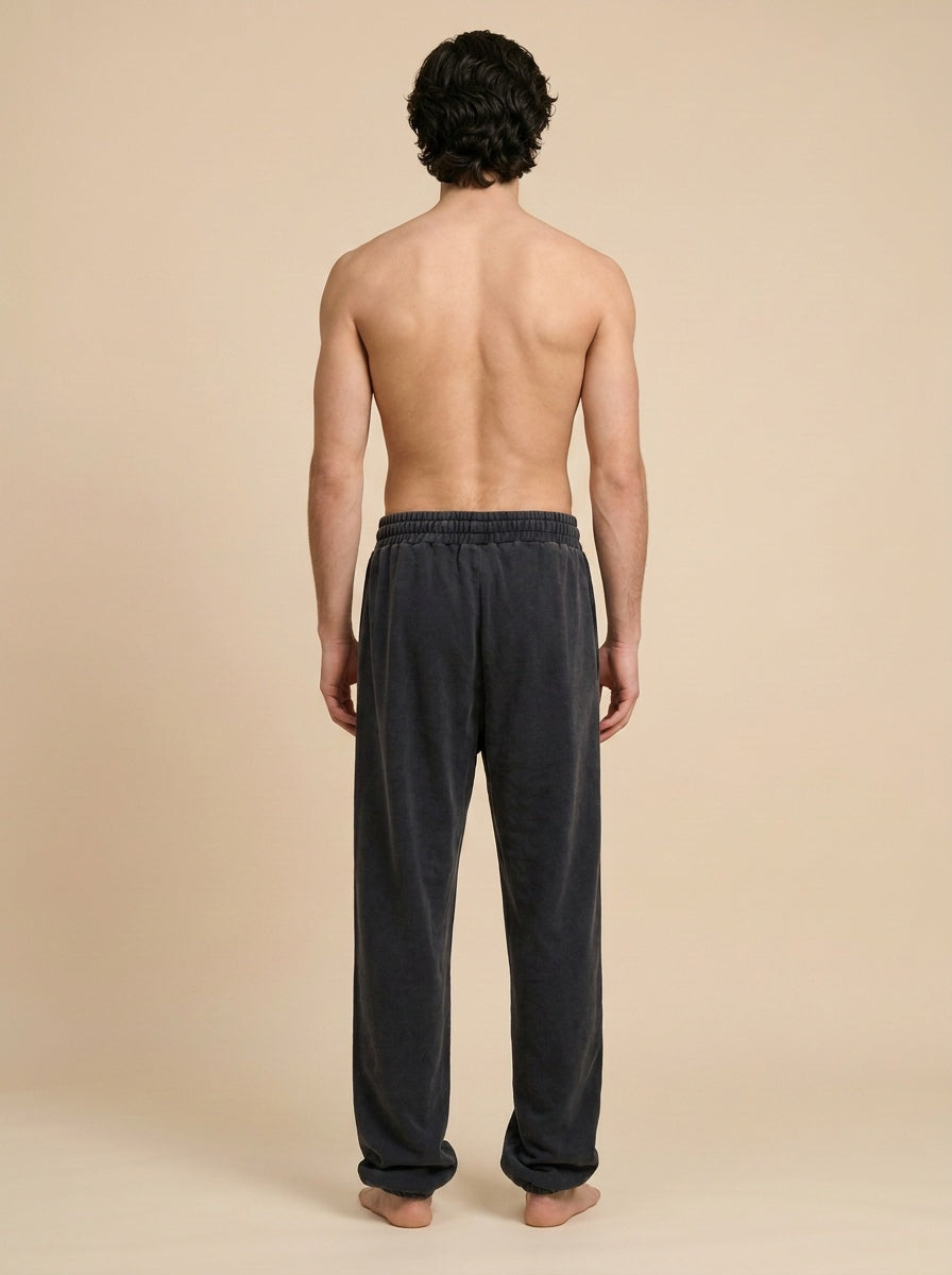 A man viewed from behind wearing dark charcoal gray lounge pants, standing against a solid beige studio background.
