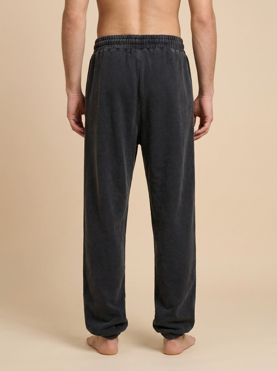 A man viewed from the back wearing dark charcoal gray lounge pants with an elastic waistband, standing barefoot against a solid beige studio background.