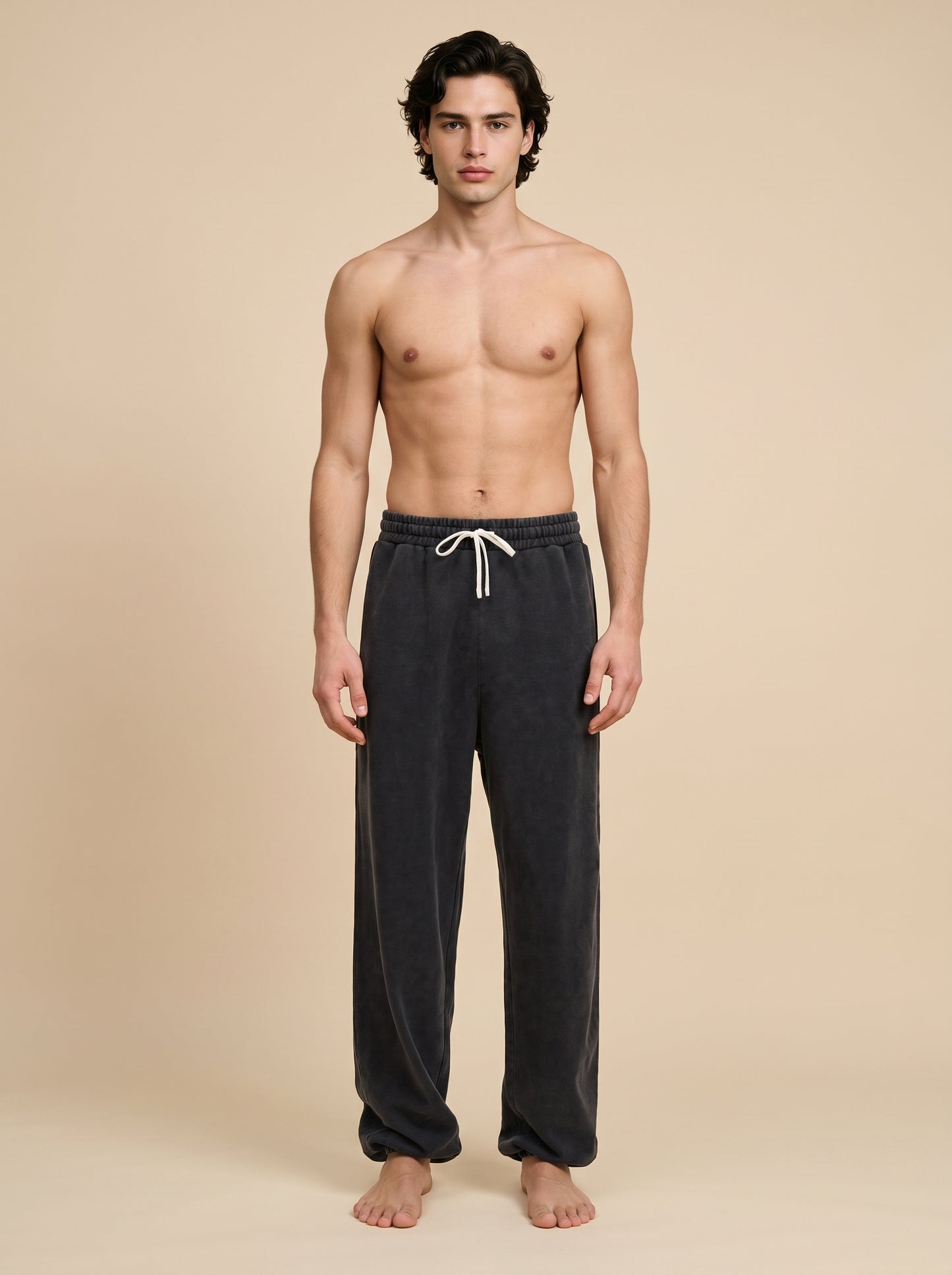 A shirtless male model stands against a plain beige studio background wearing dark charcoal gray lounge pants with a white drawstring.