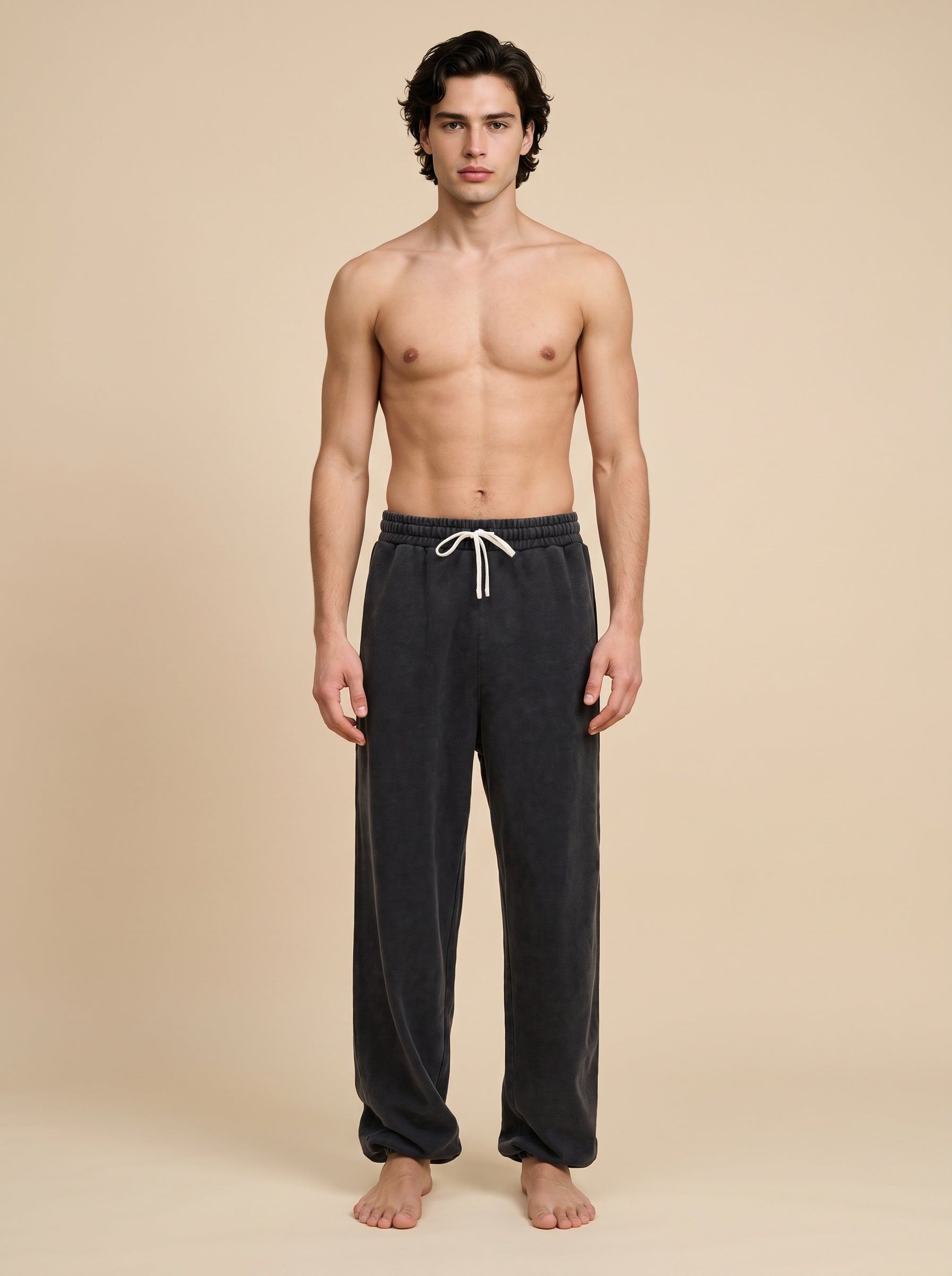 A shirtless male model stands against a plain beige studio background wearing dark charcoal gray lounge pants with a white drawstring.
