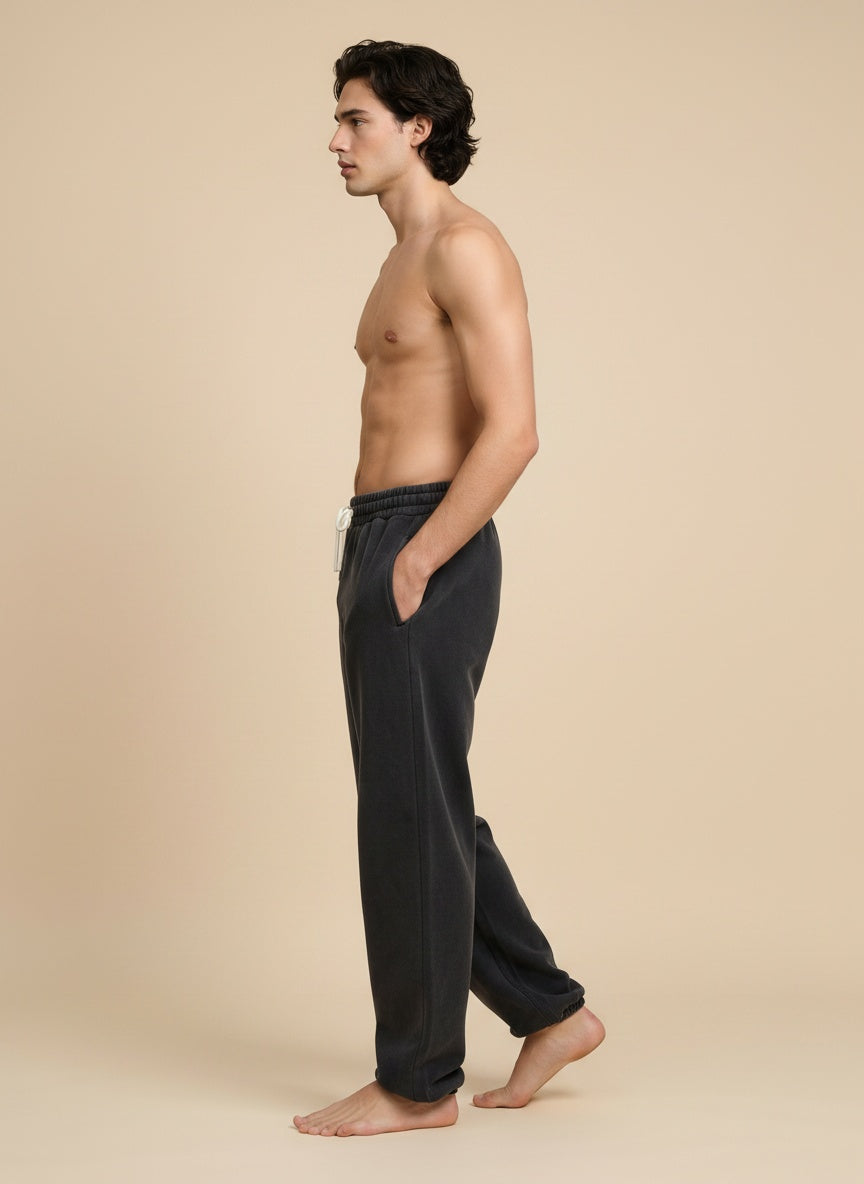 A shirtless man in profile wearing dark charcoal gray lounge pants, standing against a solid beige studio background.