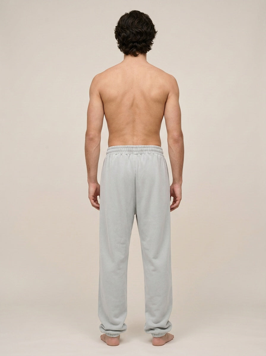 A man viewed from behind wearing light gray sweatpants, standing against a plain beige studio background.
