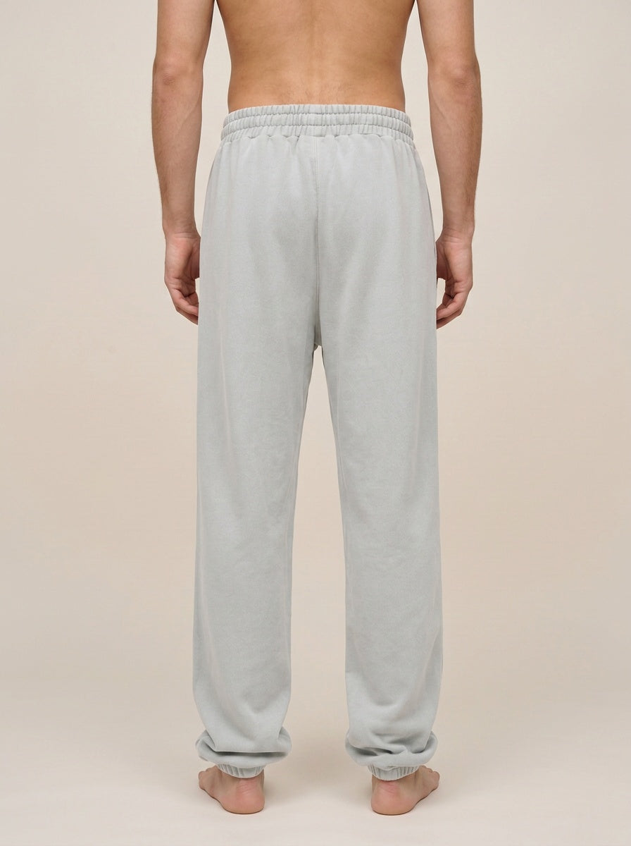 A man viewed from behind wearing light gray sweatpants, standing barefoot against a plain beige studio background.