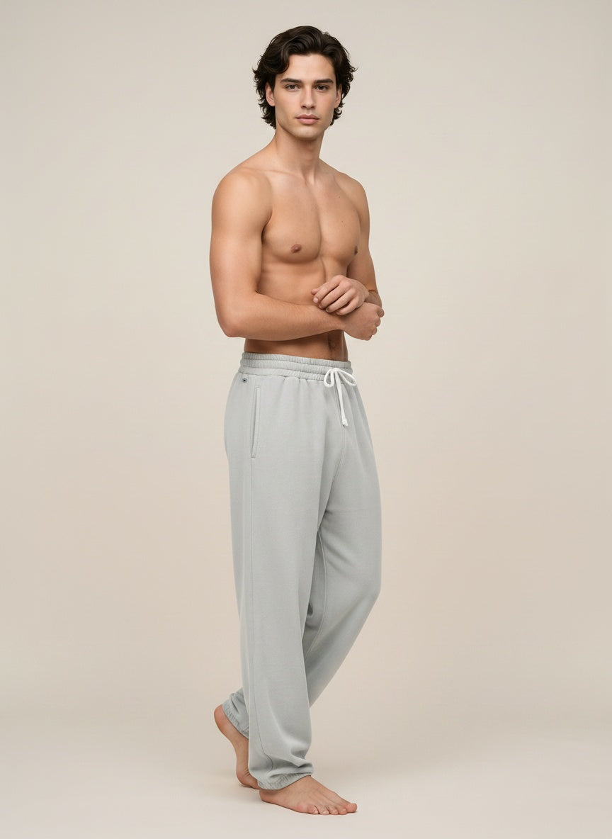 A shirtless male model wearing light gray lounge pants with a white drawstring, standing against a plain beige studio background.