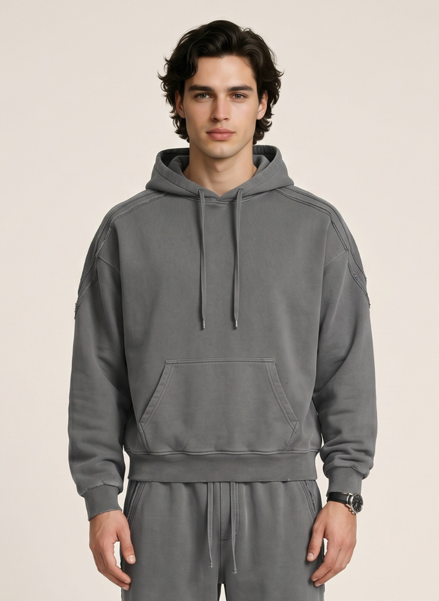 A man wearing a gray pullover hoodie with matching drawstrings and sweatpants, standing against a plain light beige studio background.