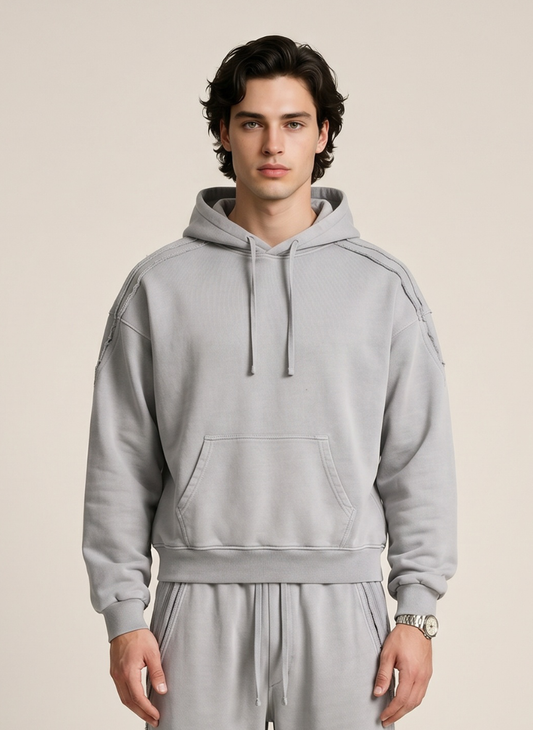 A man wearing a light gray pullover hoodie with drawstrings and a kangaroo pocket, paired with matching gray sweatpants, against a solid beige studio background.