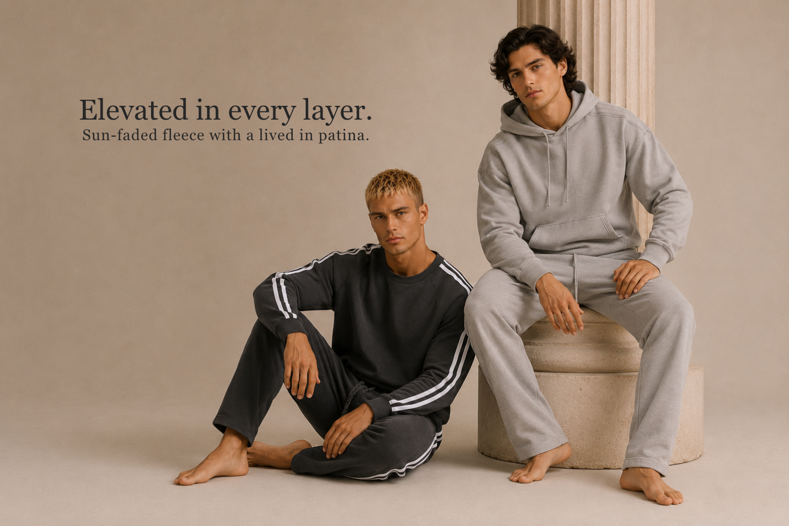 Two men modeling matching loungewear sets in a minimalist studio setting. One man sits cross-legged wearing a dark charcoal fleece sweatshirt and pants with white stripes, and the other stands on a pedestal wearing a light gray fleece hoodie and sweatpants.