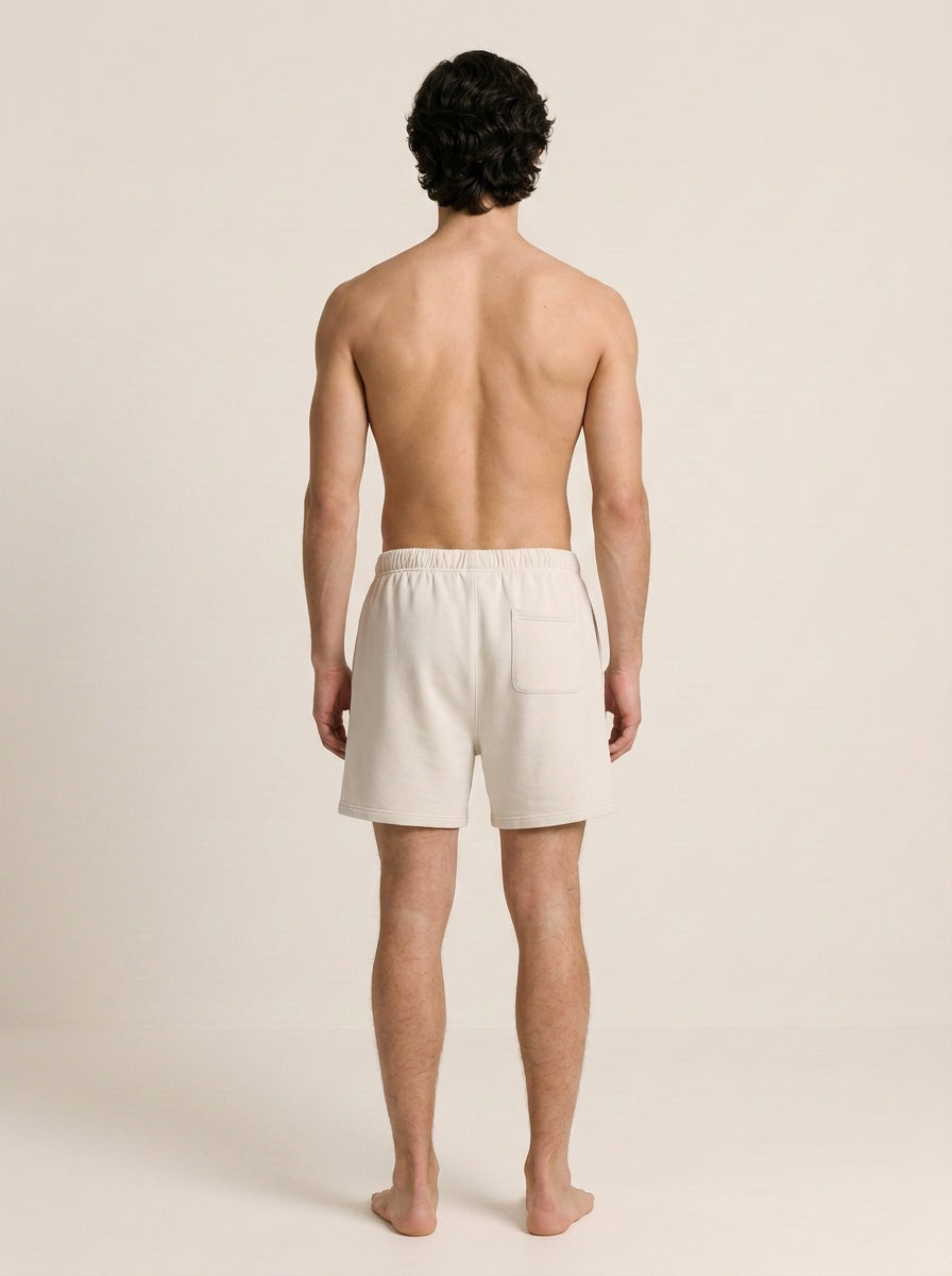 A man viewed from behind wearing light cream-colored drawstring shorts, standing against a solid light beige studio background.